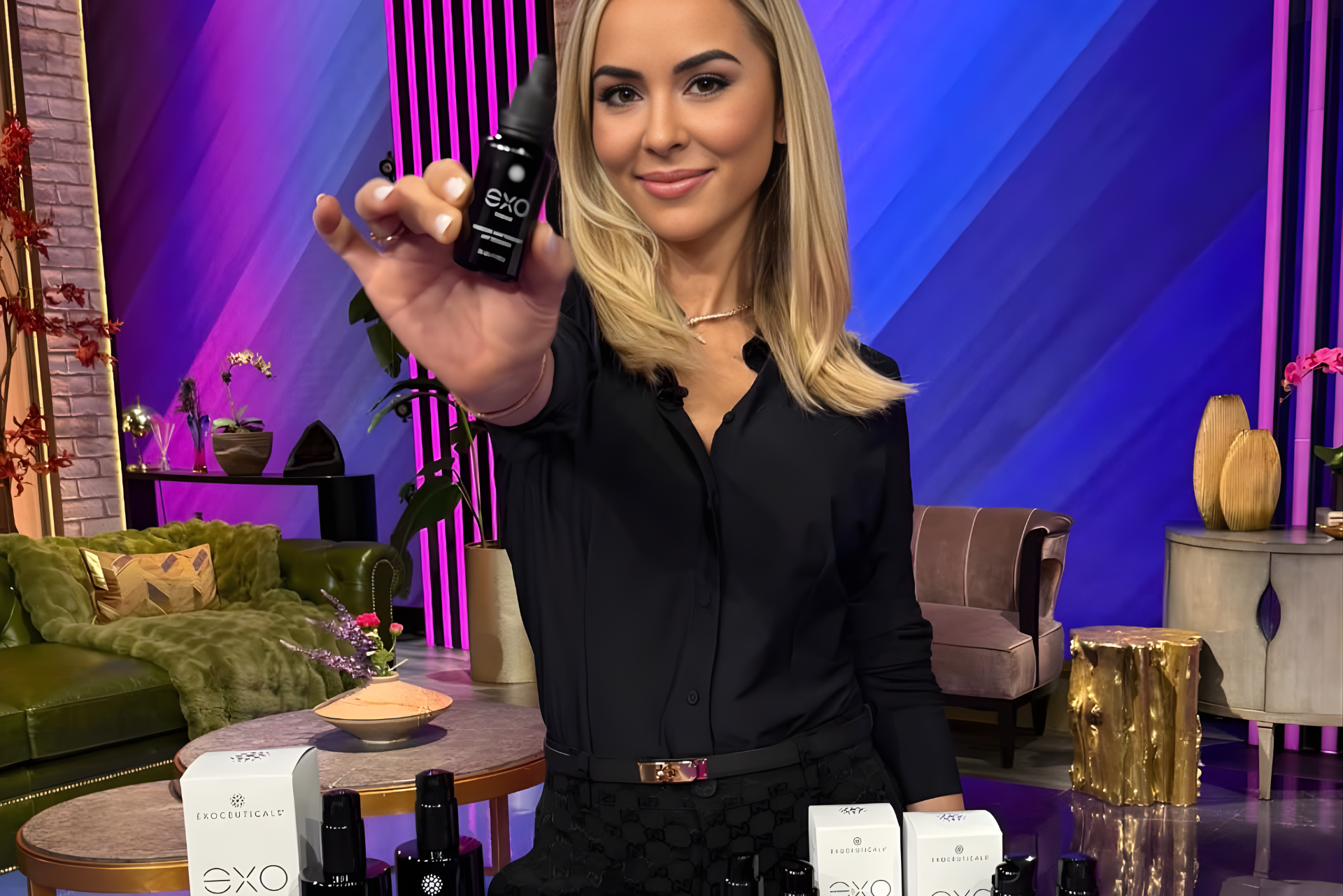 Woman holding a black product bottle, smiling, on a set with products and furniture.