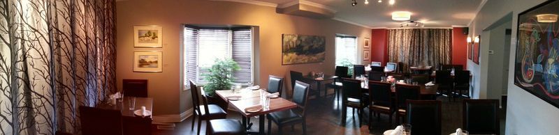 A restaurant dining room with tables set, curtains, artwork, and a neutral color scheme.