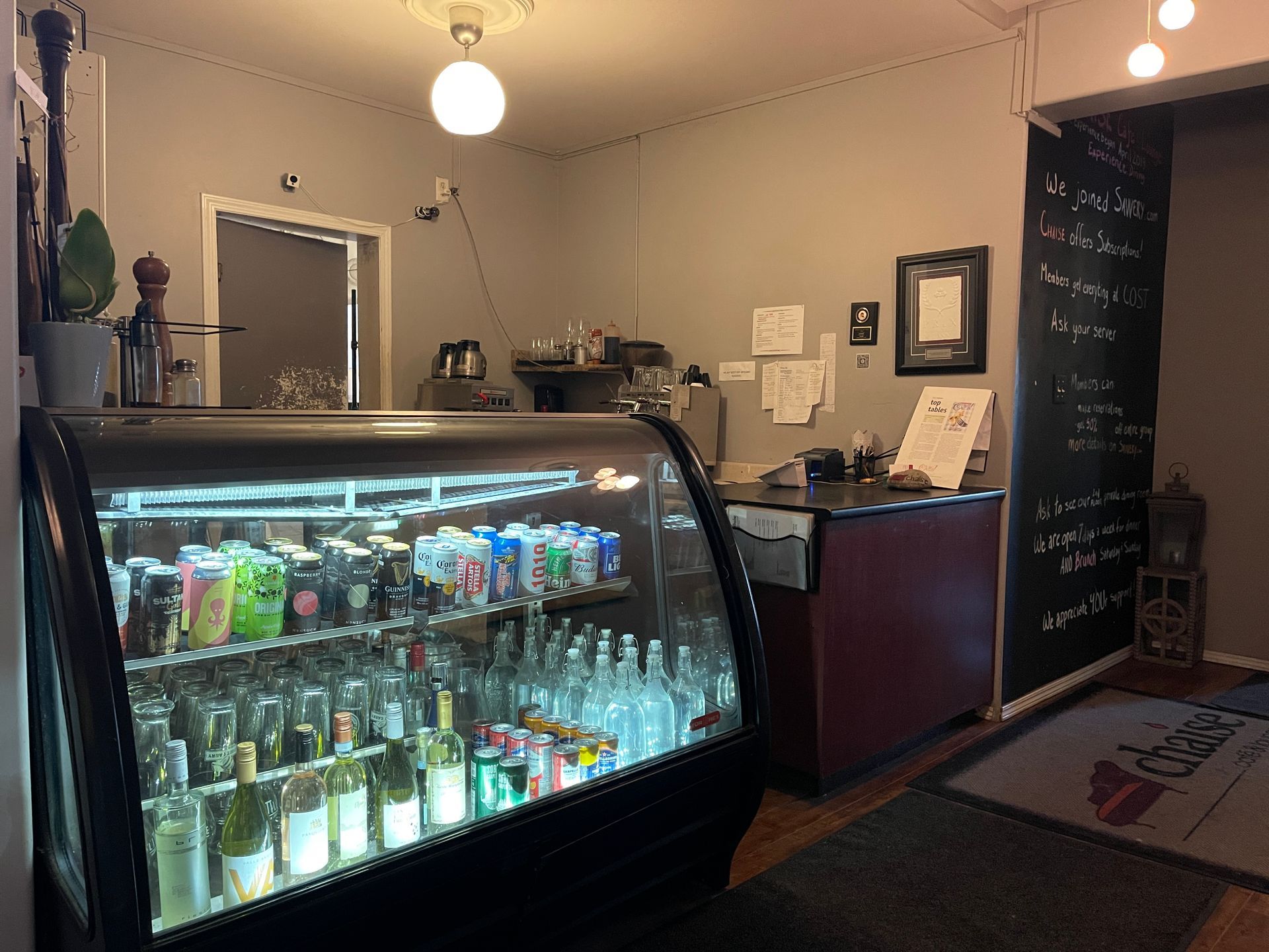Restaurant interior with bar, stools, tables, chairs, and hanging lights. Green foliage in foreground.