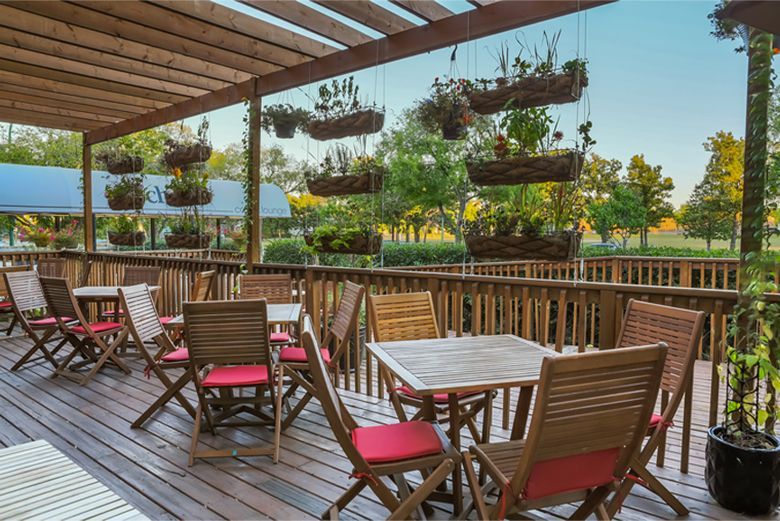 Wooden patio with tables, chairs, hanging flower baskets, and trees in the background.