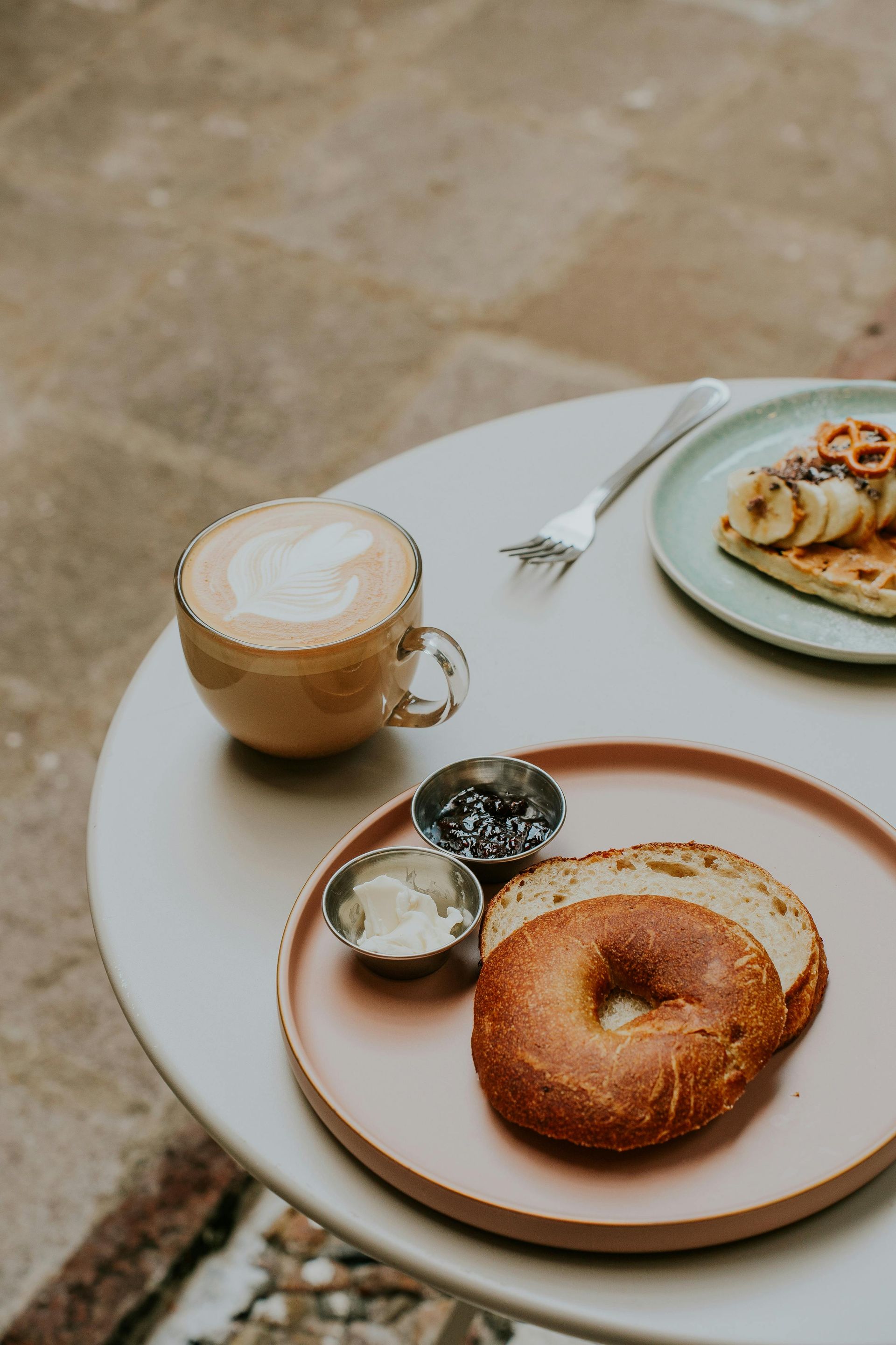 A bagel with toppings, coffee, and waffles on a cafe table.
