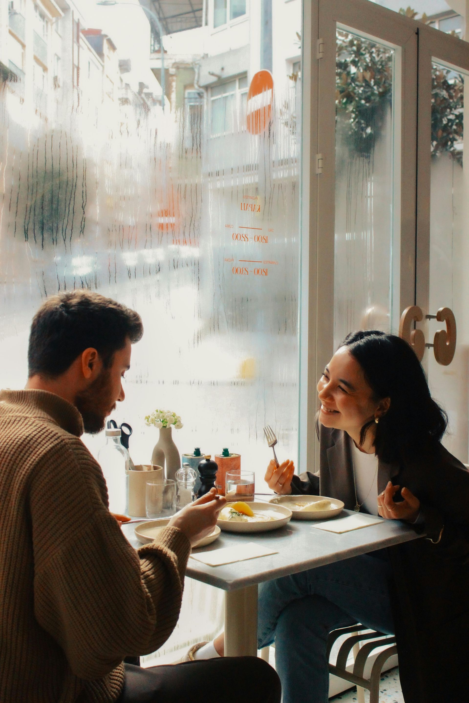 Couple smiling while eating breakfast at a cafe table near a foggy window.