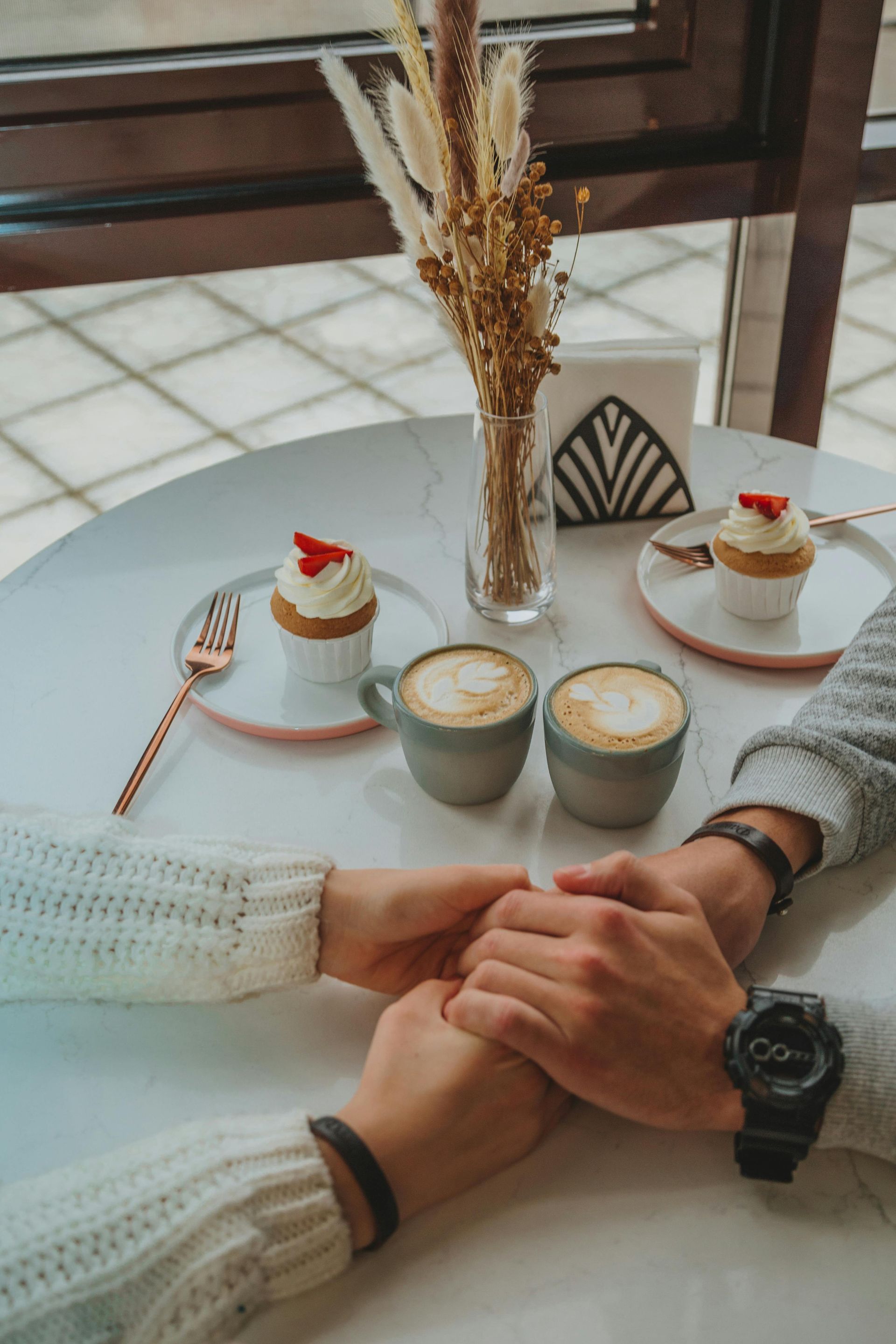 Couple holding hands over a table with coffee, cupcakes, and flowers. Cafe setting, warm colors.