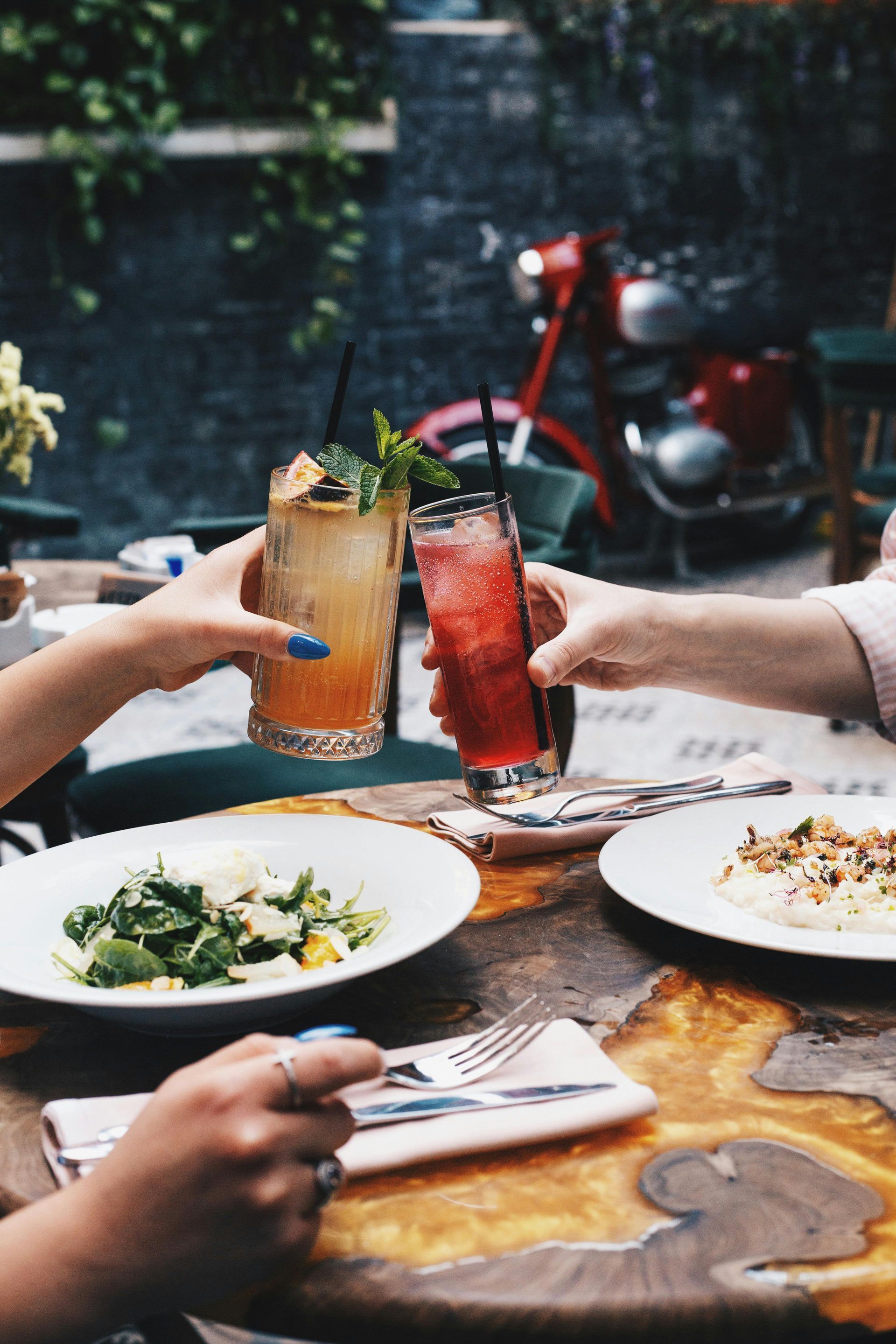 Two people toasting cocktails over food at an outdoor table with a red motorcycle in the background.