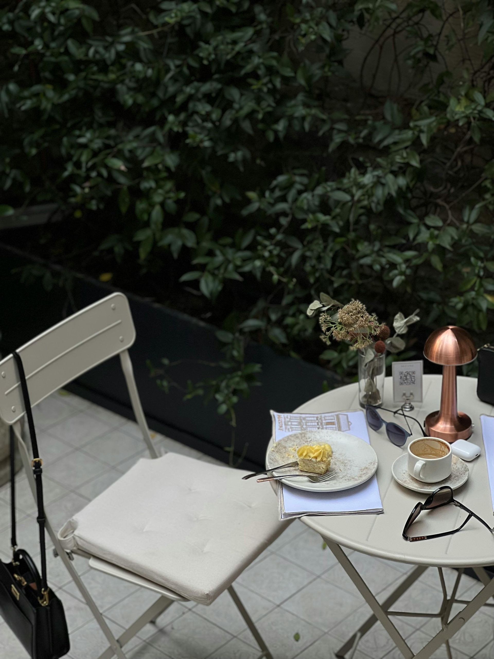A table with coffee and cake outdoors, next to a chair, near greenery.