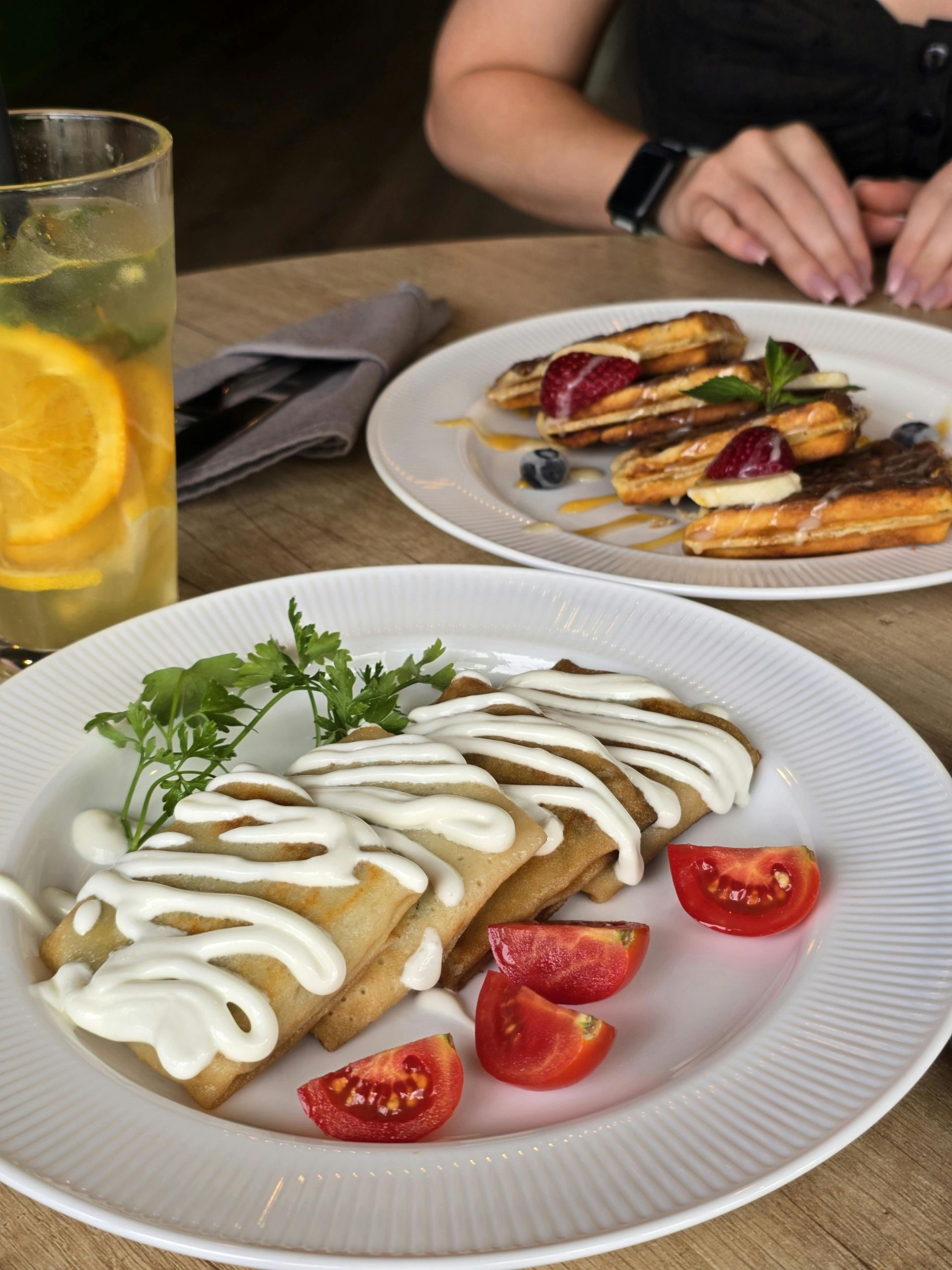 Plates of pancakes with sour cream and tomatoes, and pastries with berries on a table, along with a drink.