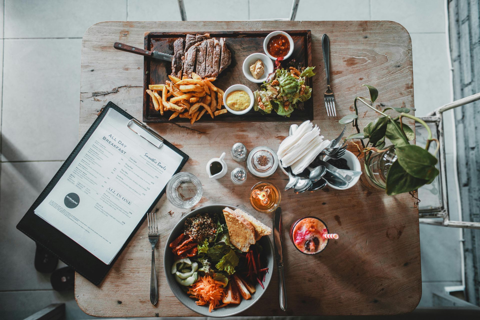 Overhead shot of a wooden table with steak, fries, salad, sauces, a menu, drink, and assorted food items.