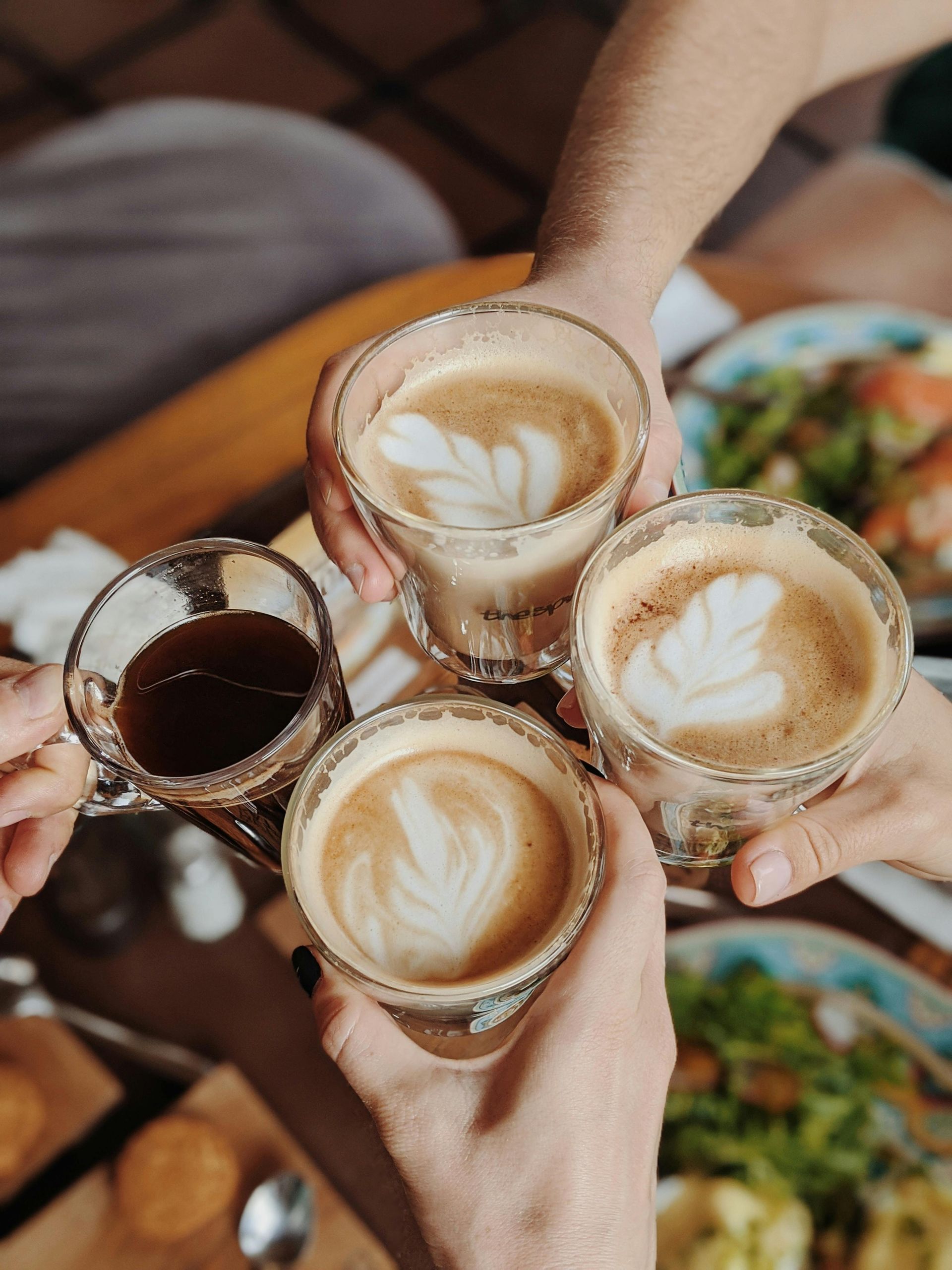 Four hands holding coffee drinks, including lattes, in a cafe setting, toasting each other.