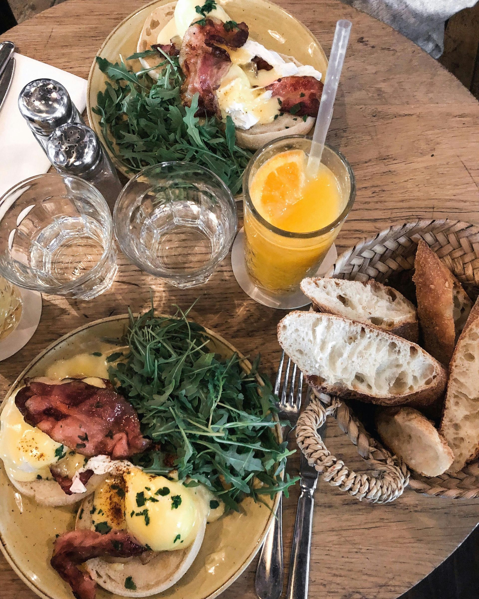 Two plates of eggs benedict with bacon, arugula, orange juice, and bread basket on a wooden table.