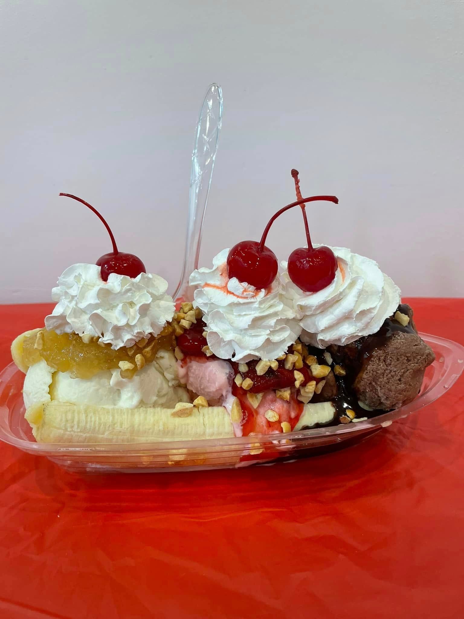 A plastic container filled with ice cream , whipped cream and cherries on a red table.