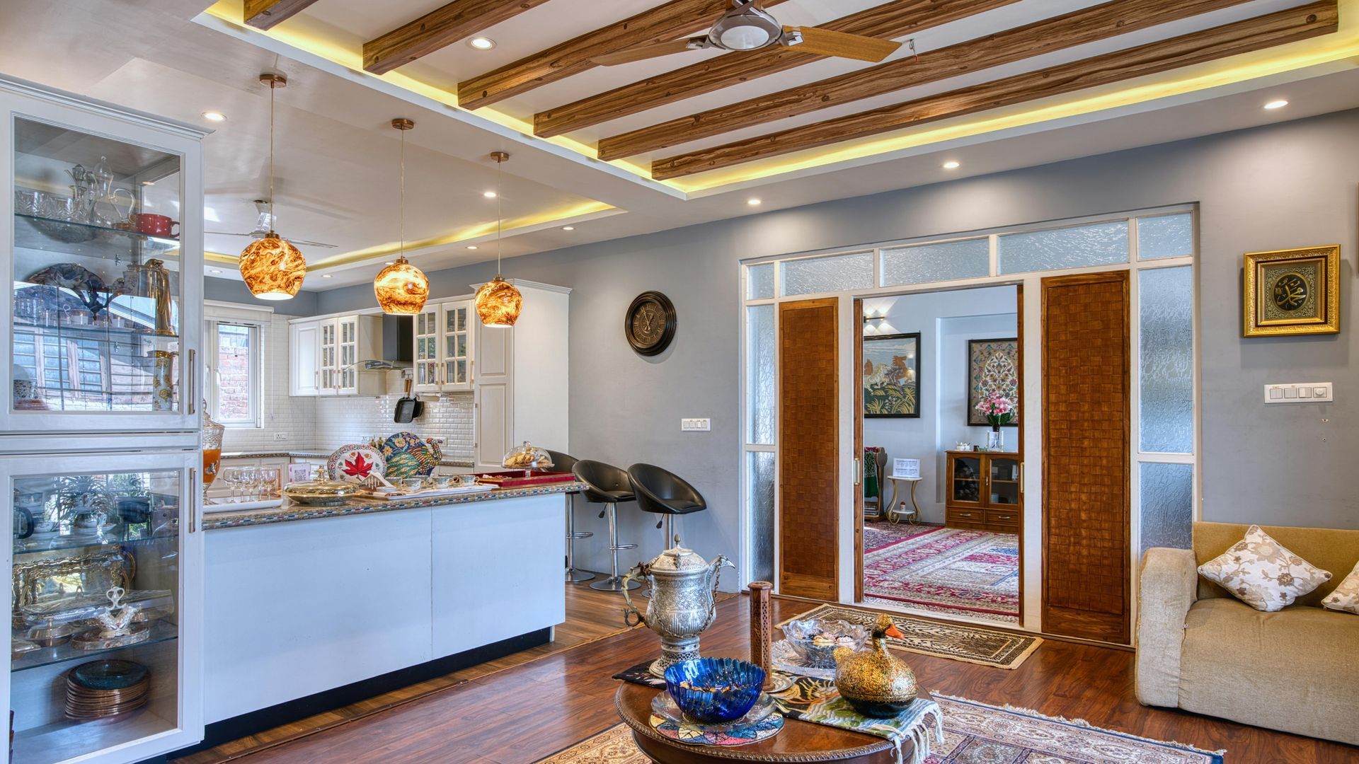 Living room with kitchen, bar, and doorway; wooden beams, white cabinets, and gray walls.