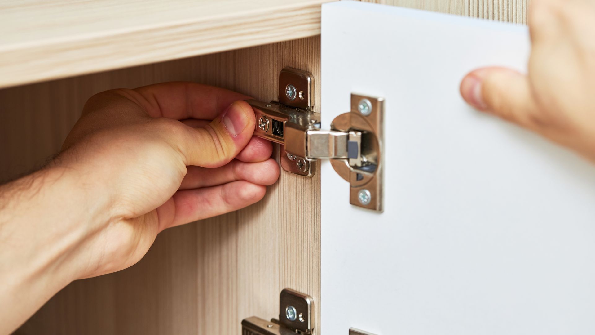 Hands adjusting a cabinet door hinge, attached to a wooden cabinet.