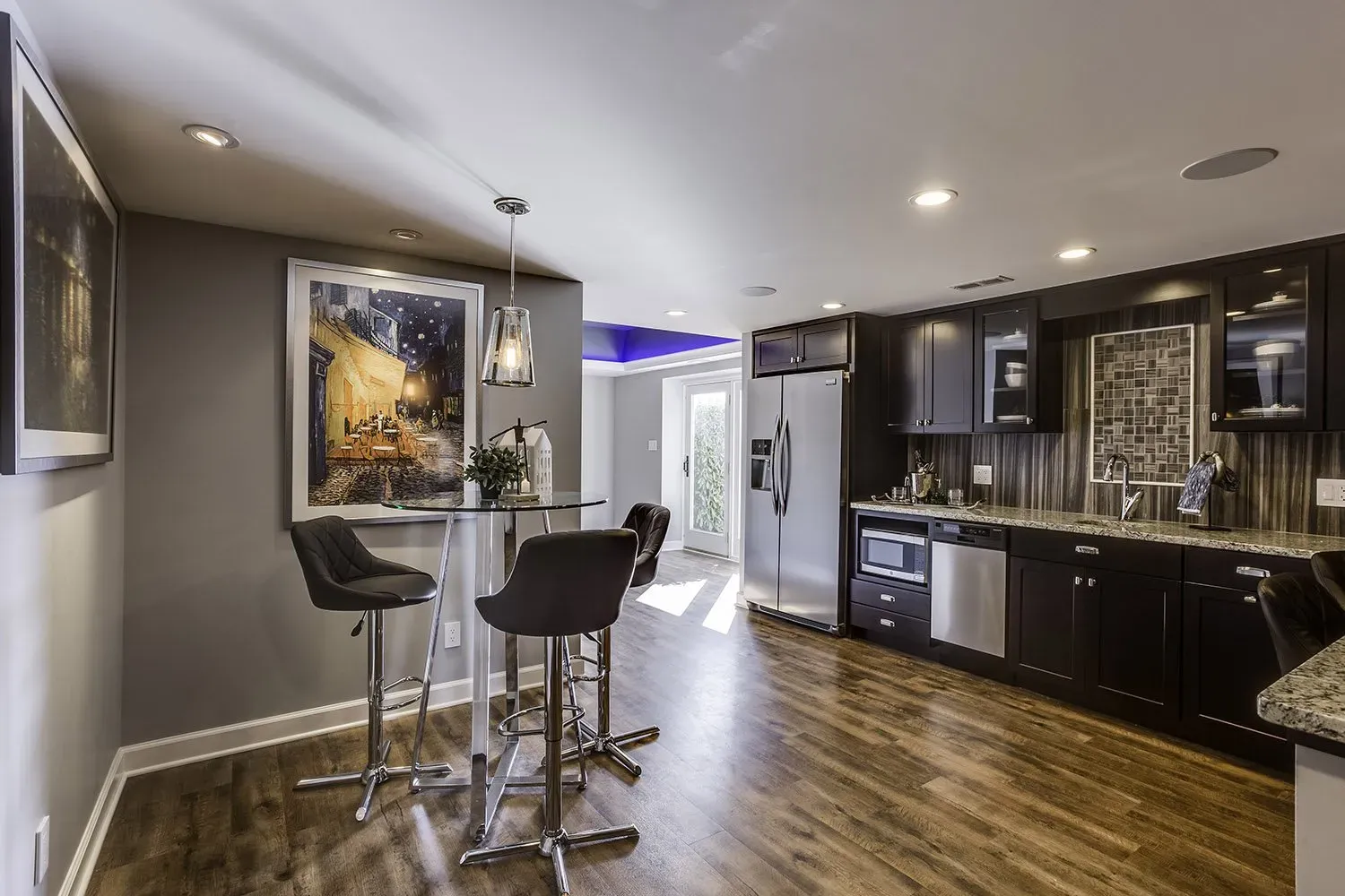 A kitchen with wooden floors , black cabinets , stainless steel appliances and a bar.