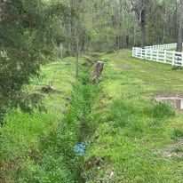 A stream running through a lush green field with a white fence in the background.