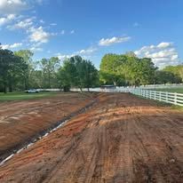 A dirt road with a white fence and trees on the side.
