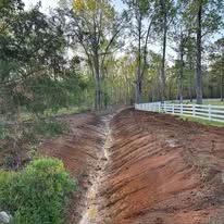 A dirt road leading to a white fence surrounded by trees.