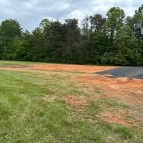 A baseball field with trees in the background and a dirt road in the foreground.