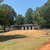 A house is sitting on top of a dirt hill in the middle of a field surrounded by trees.
