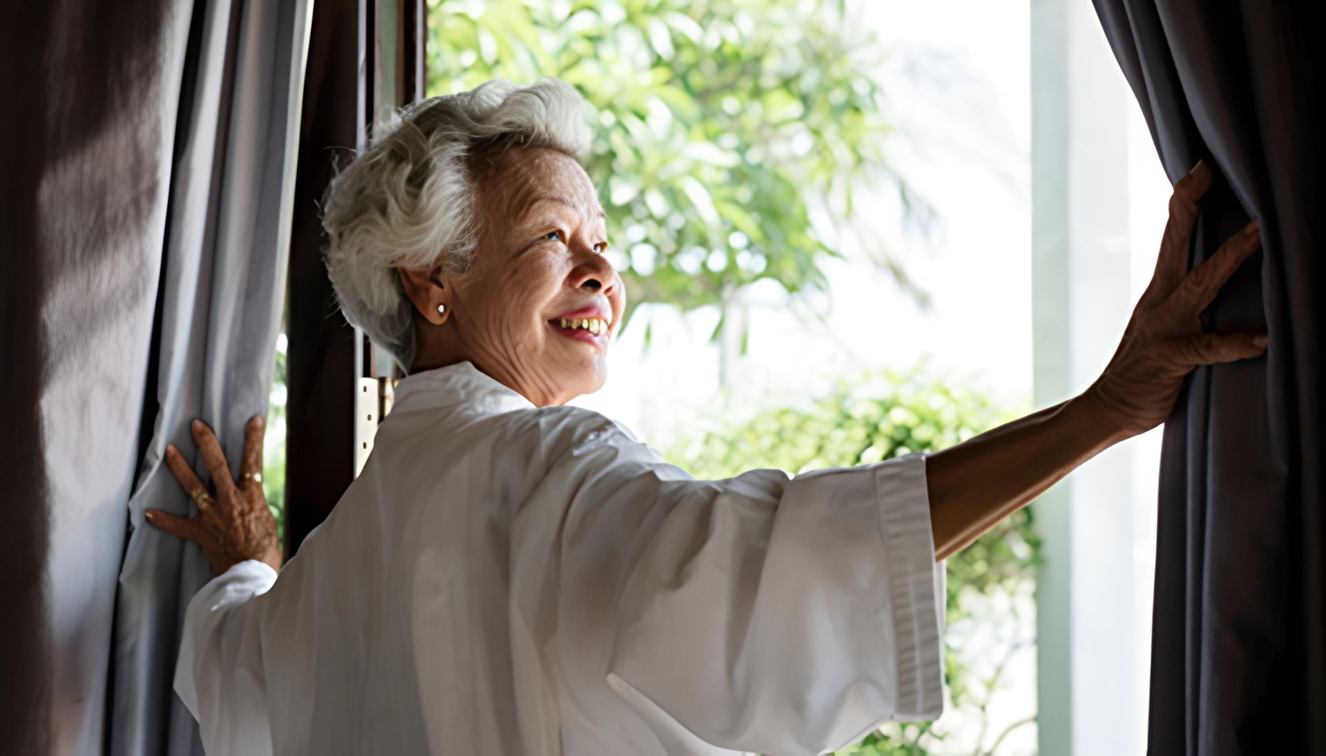 Woman in white robe, smiling, opening curtains to look outside.