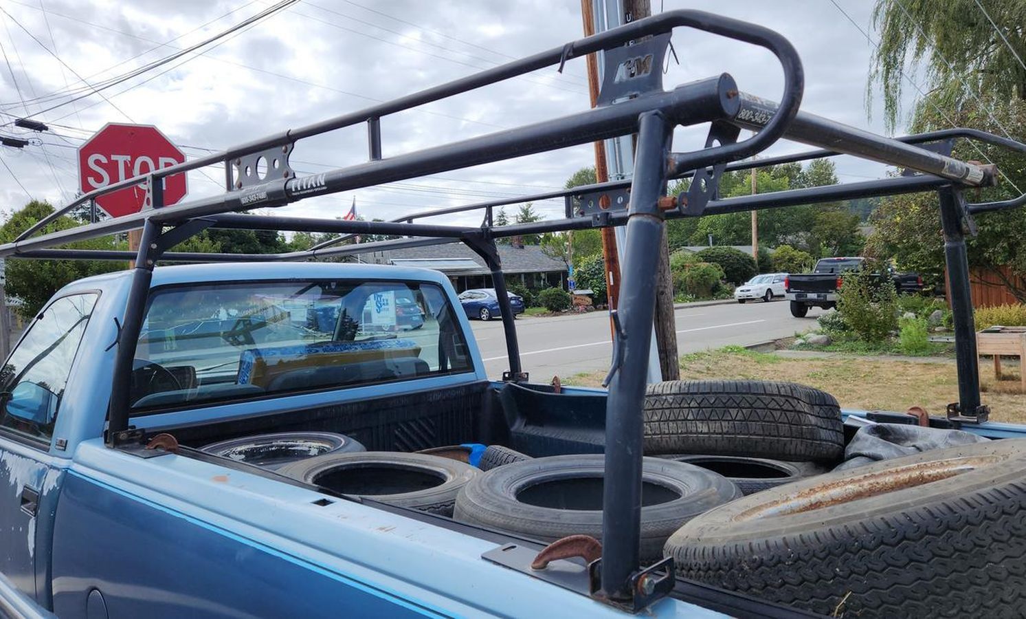 A blue truck with a roof rack and tires in the back is parked in front of a stop sign.