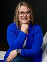 A woman wearing glasses and a blue jacket is sitting on a bed.