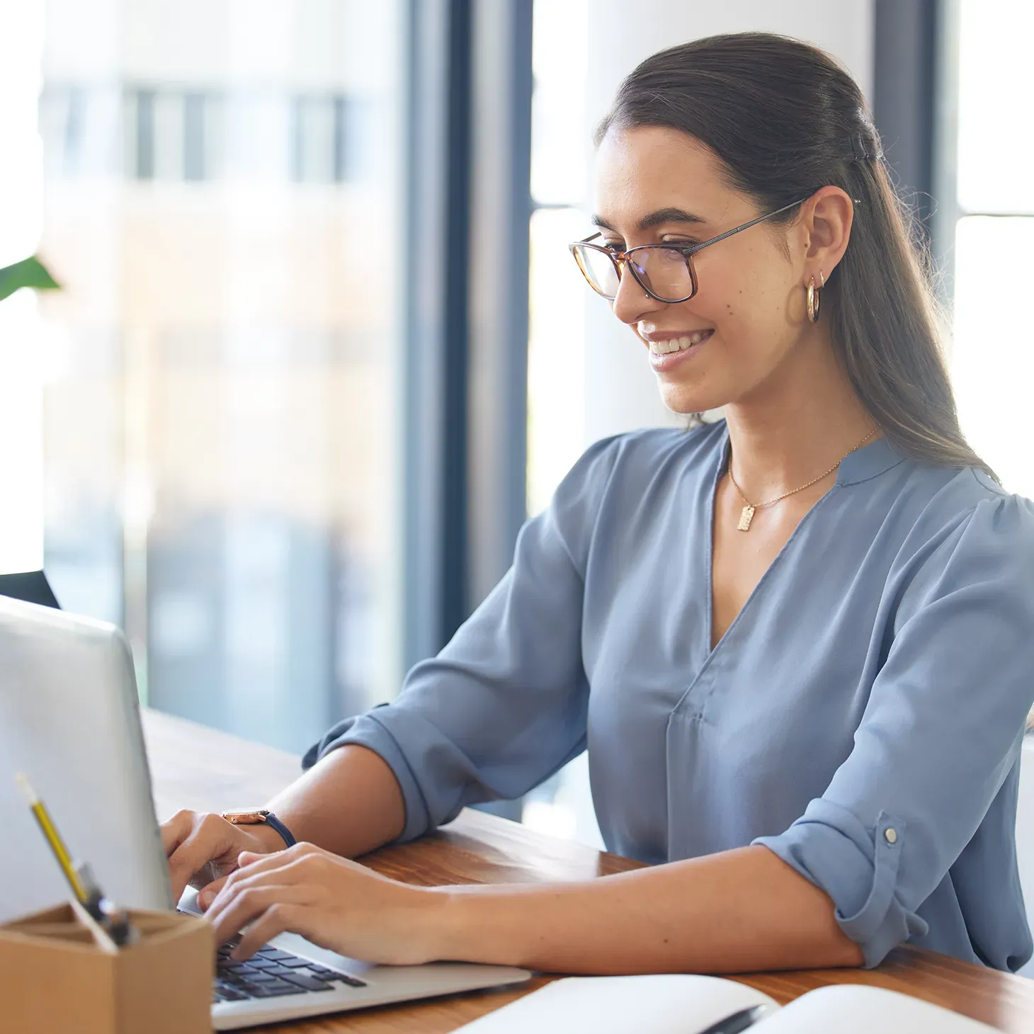 Woman with glasses smiles while typing on a laptop at a wooden desk in a bright office.