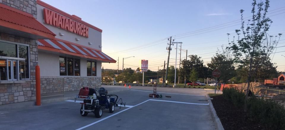 A worker operates a white line-striping machine in the parking lot of a Whataburger restaurant during the day.