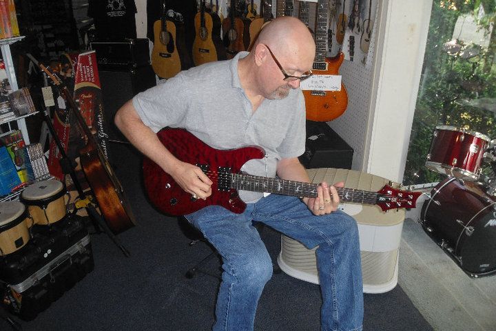 Man seated playing a red electric guitar in a music store, surrounded by hanging guitars.