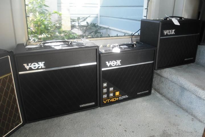 Three VOX guitar amplifiers on a porch, including two black amps and one smaller black amp on steps