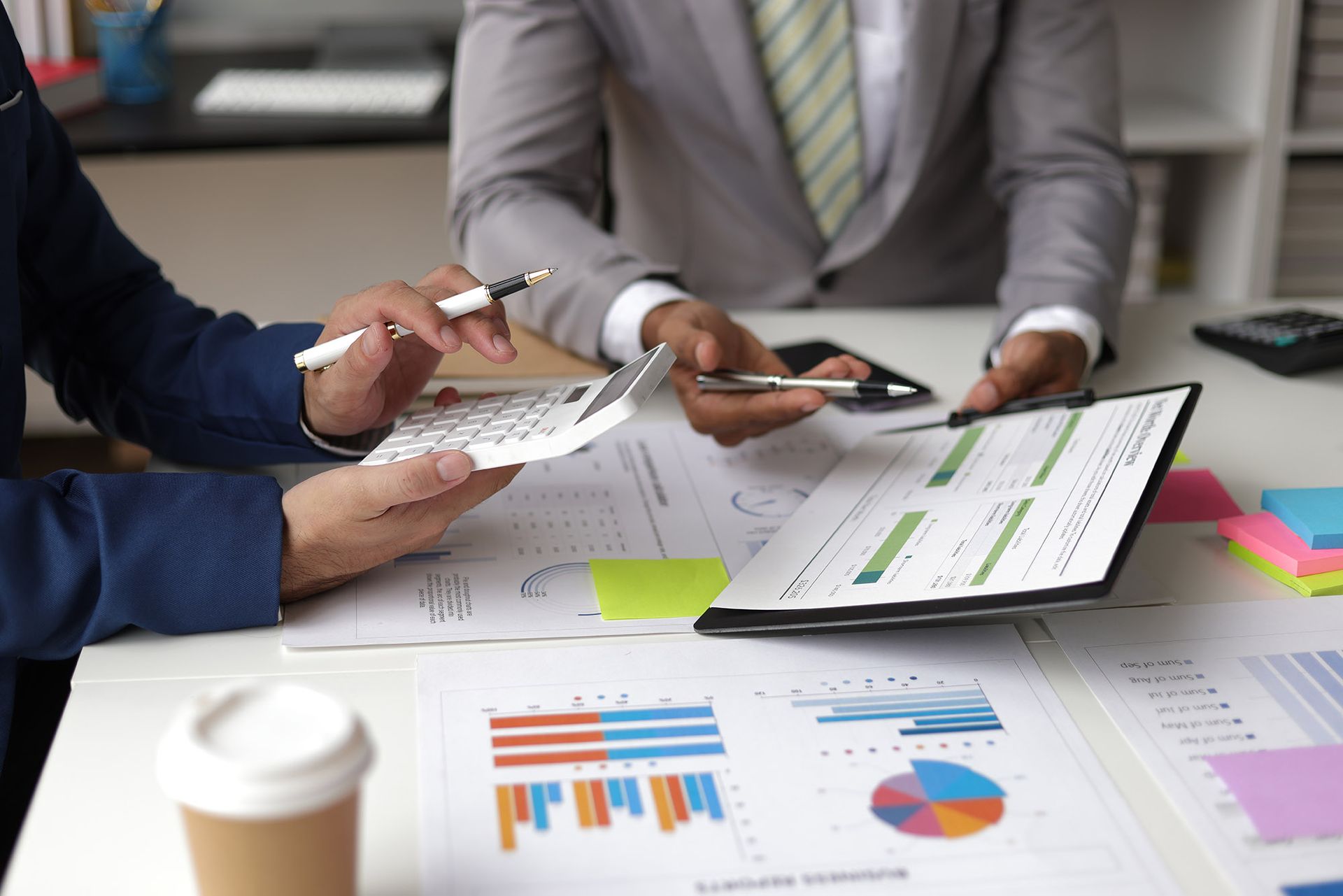Two people in suits reviewing charts and using a calculator at a desk, office setting.