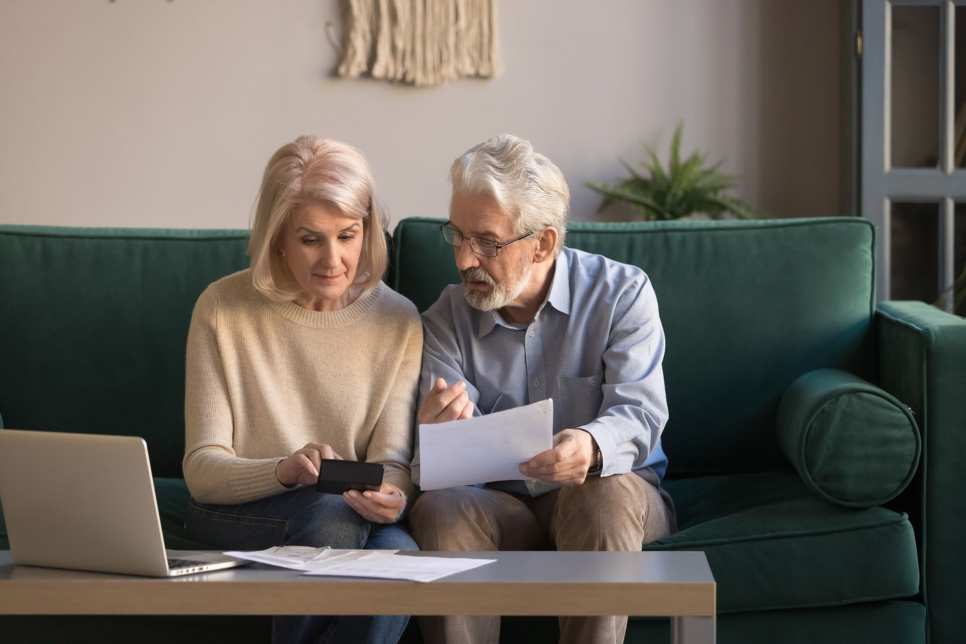 Older couple reviews finances on a laptop and paper, sitting on a green couch.