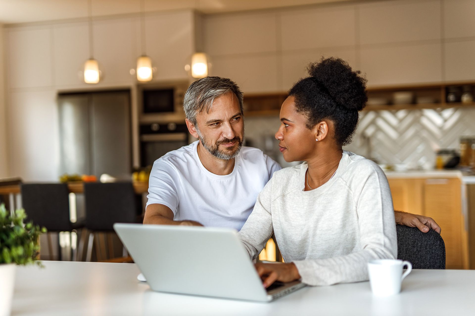 Couple at a kitchen table looking at a laptop. The woman is typing. Man has his arm around her shoulder.