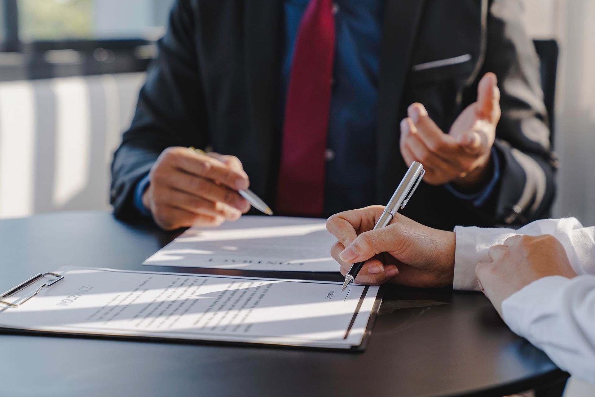 Person signing a document, seated across from a person in a suit, both at a table.