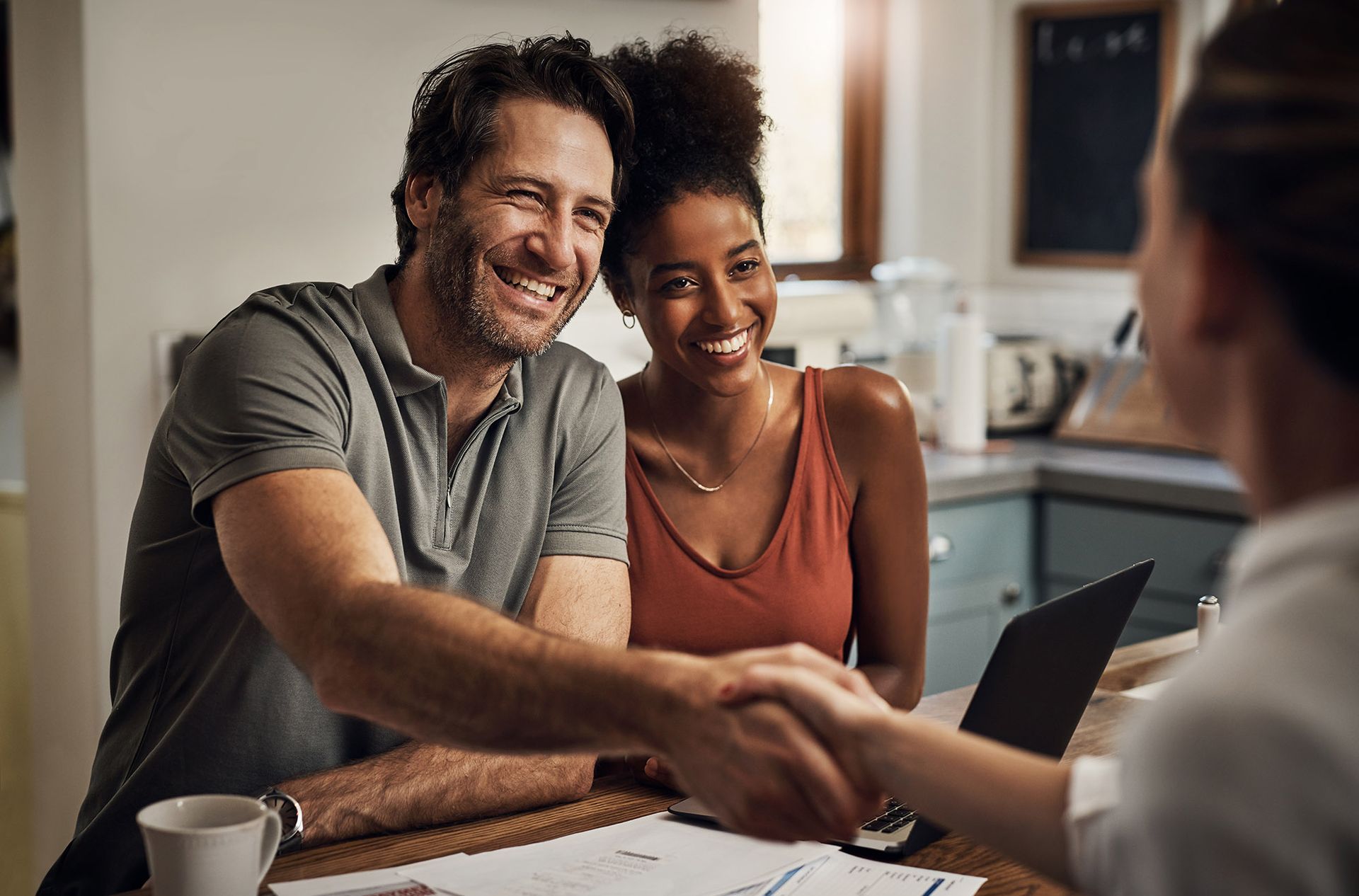 Couple shaking hands with a person, smiling, over a table with a laptop, in a kitchen.