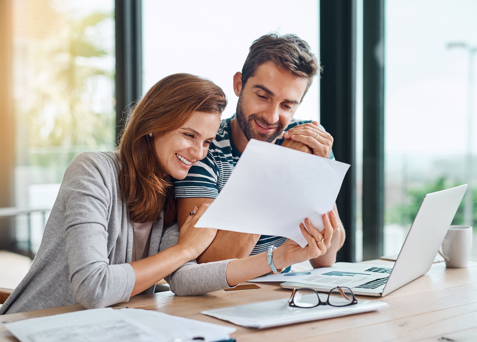 Couple smiling, reviewing paperwork at a table with laptop, glasses, and coffee cup; indoor setting.