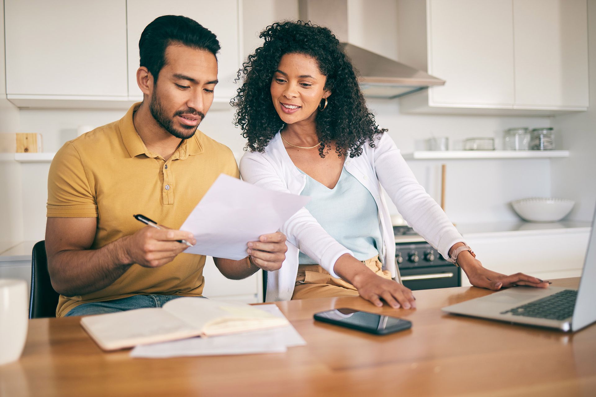 Couple reviewing documents at a kitchen table. Man holds pen, woman points. Laptop and phone nearby.