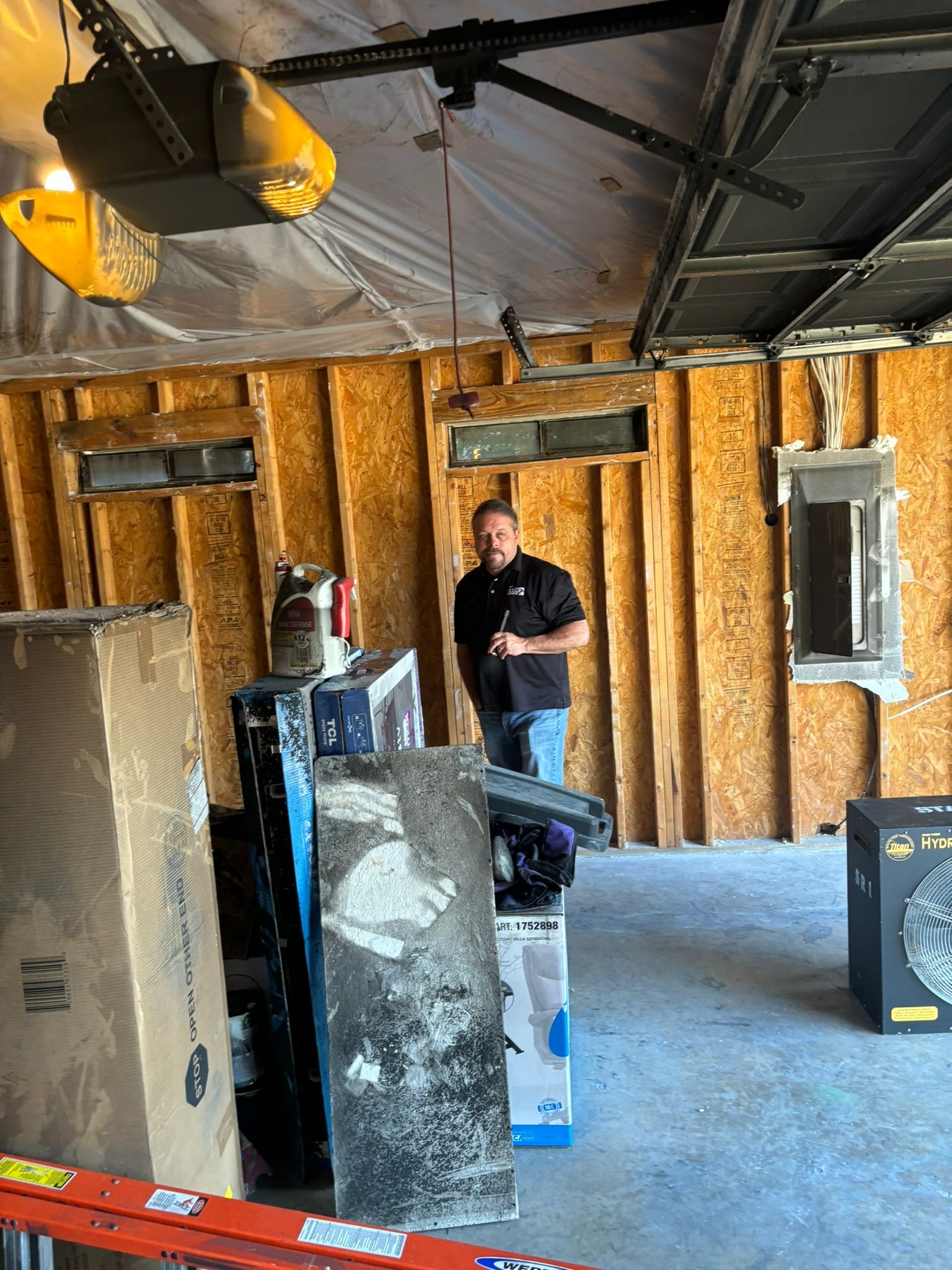 Man in garage stands amidst building materials, looking at the camera. Garage interior shows exposed studs.
