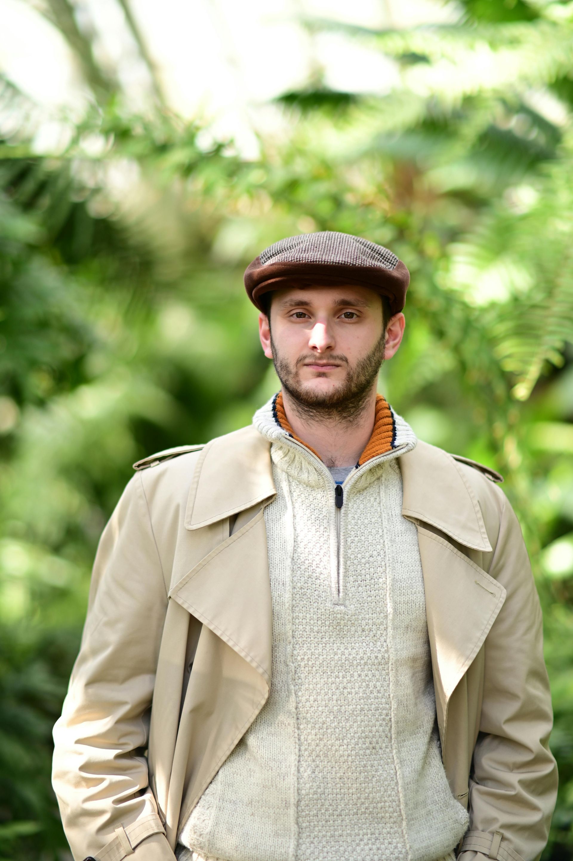 Man in cap and trench coat stands in front of green foliage, hands in pockets, looking forward.