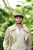 Man in cap and trench coat stands in front of green foliage, hands in pockets, looking forward.