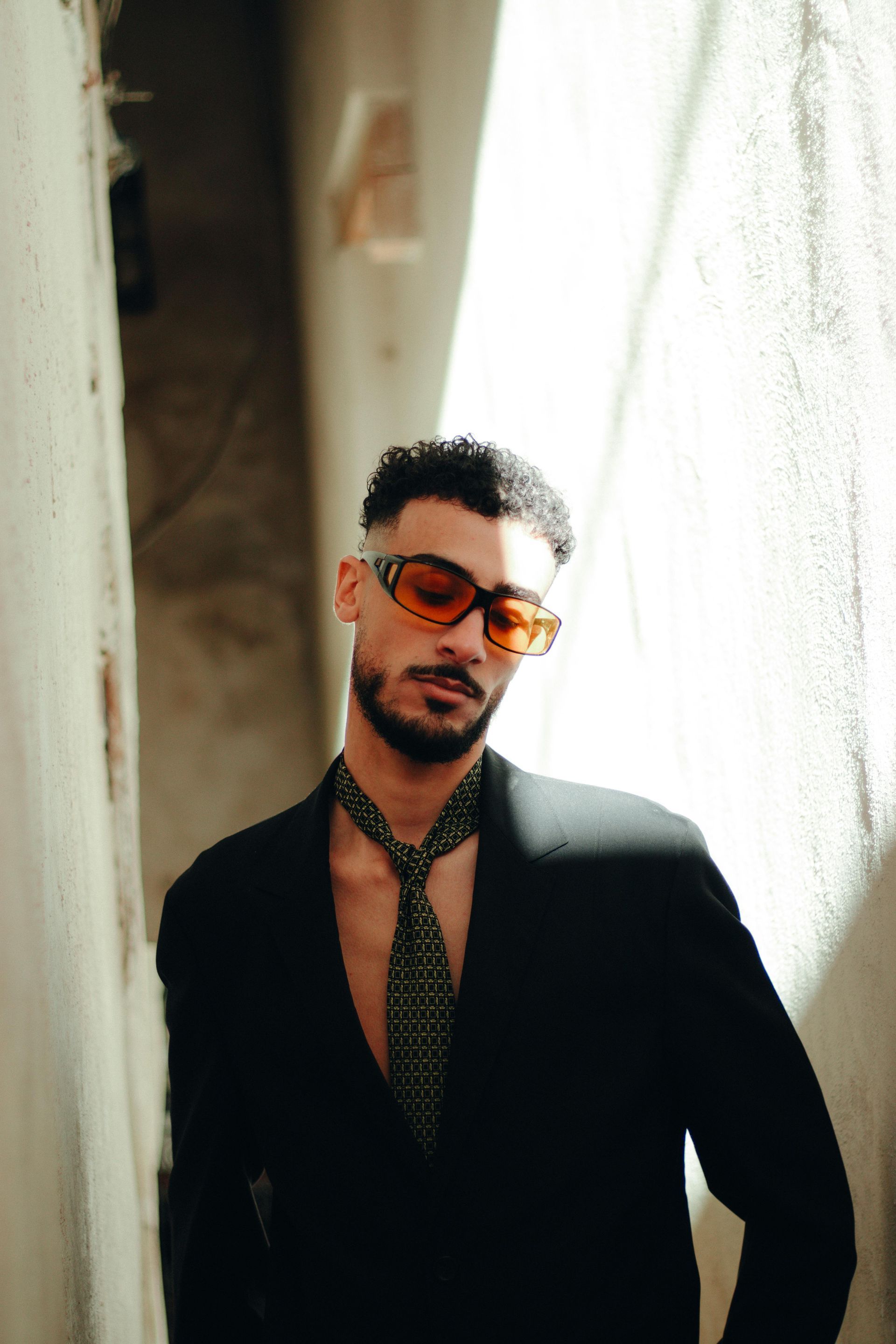 Man wearing sunglasses and blazer, standing in a building, leaning.
