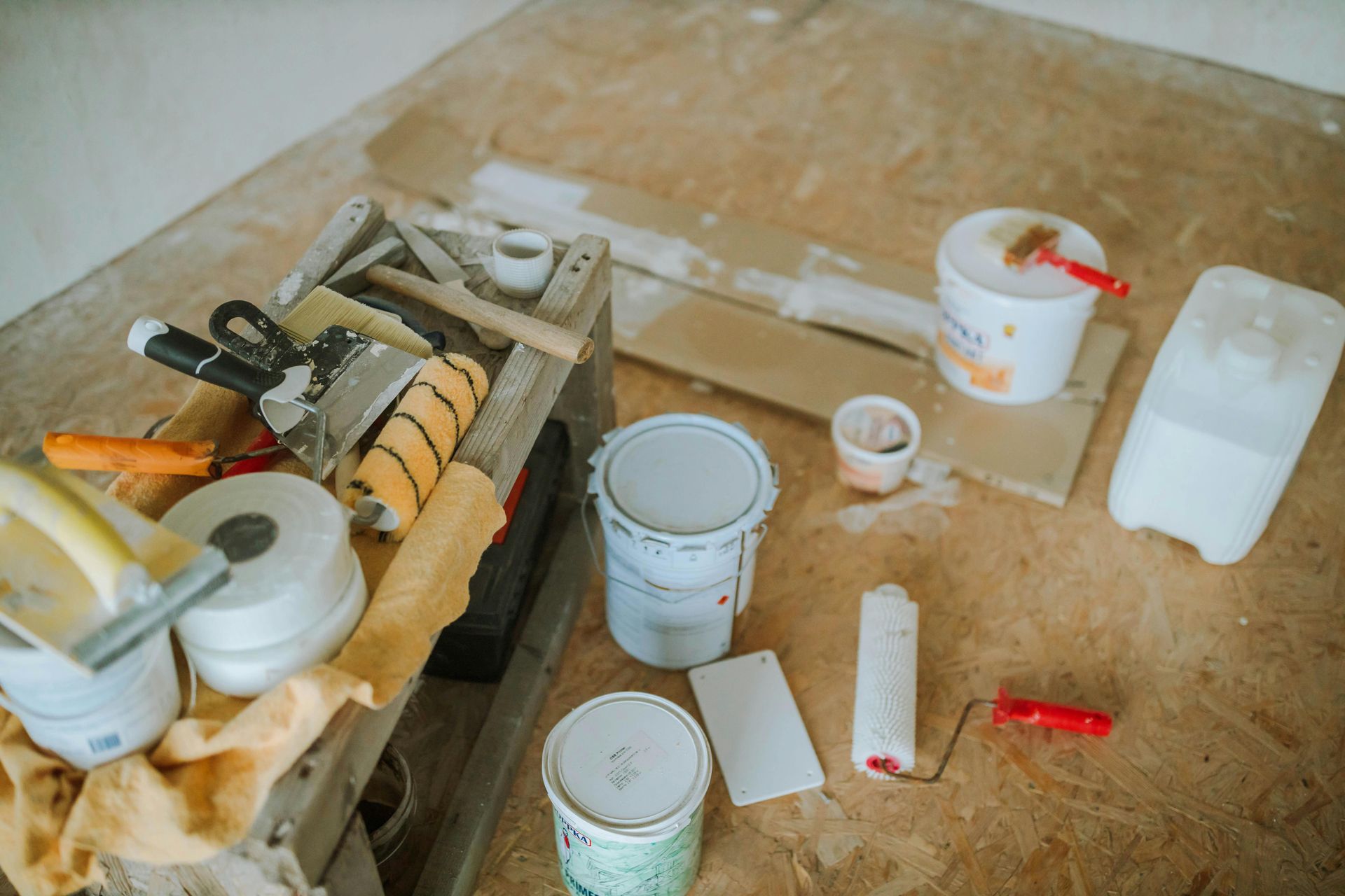Painting supplies on a wooden floor: paint cans, rollers, brushes, putty knives.