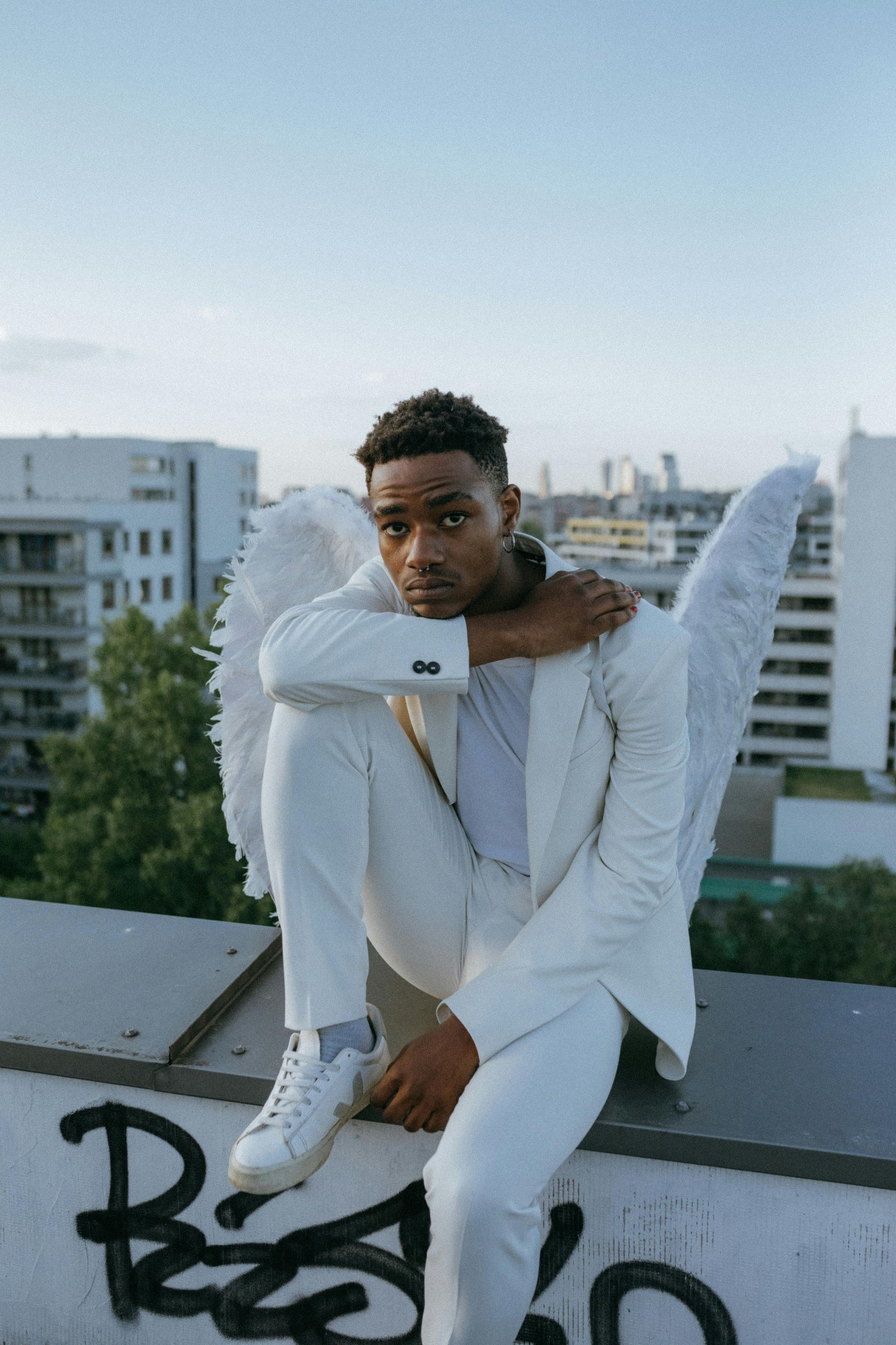 Man in white suit with angel wings sits on a rooftop, city in background.