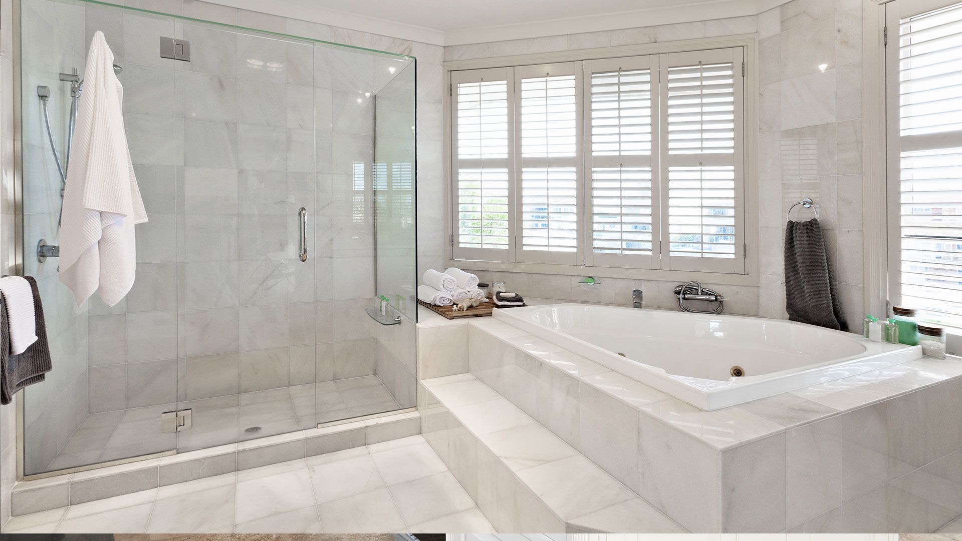 Luxurious white marble bathroom with glass shower, soaking tub, and shuttered windows.
