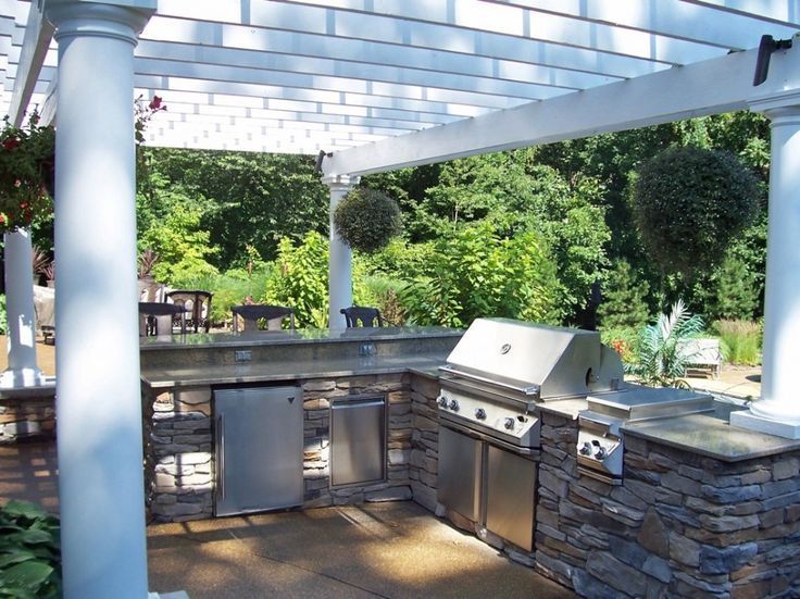 Outdoor stone patio kitchen with built-in grill and sink under a white pergola, surrounded by greenery