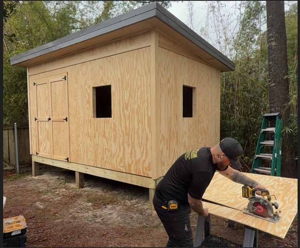 Worker cutting plywood beside a small unfinished wooden shed in the woods