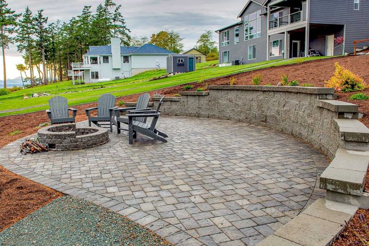 Stone patio with fire pit and chairs beside a terraced backyard retaining wall and steps