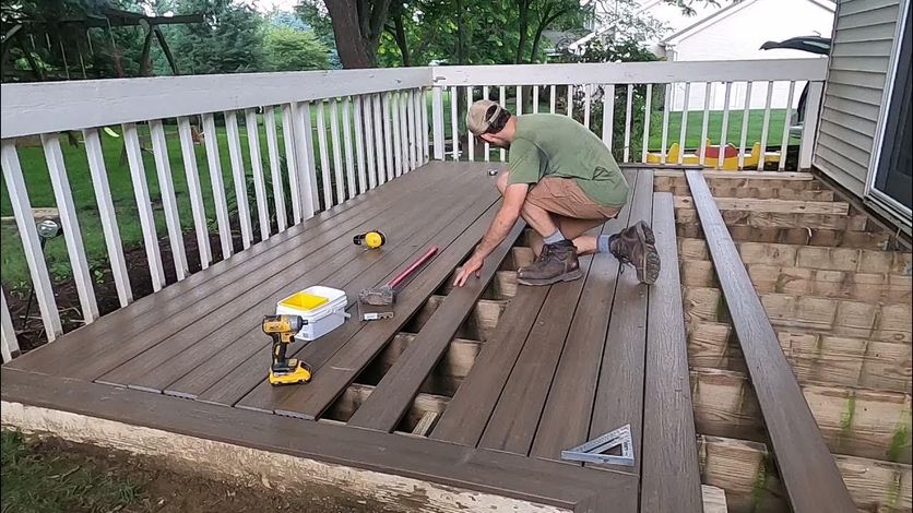 Person kneeling on a porch deck, measuring and aligning brown deck boards with tools nearby.