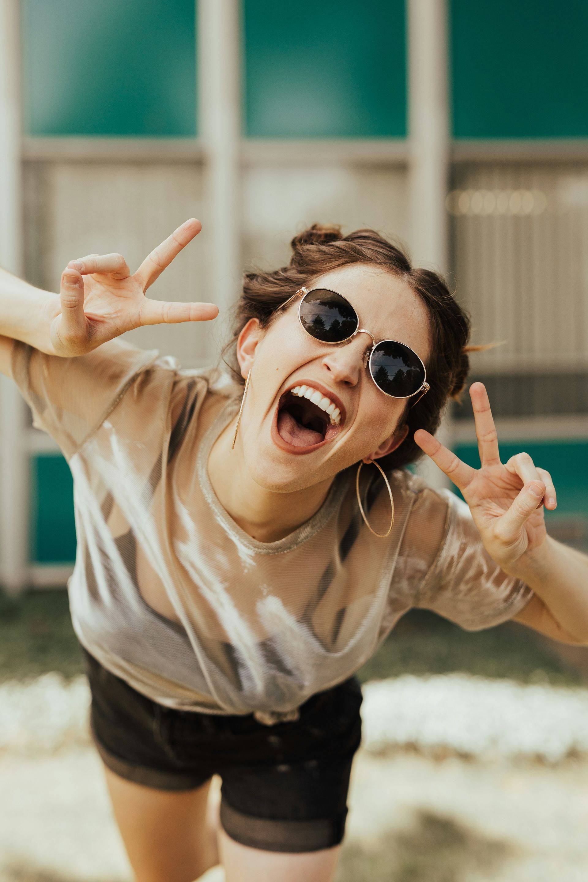 Woman with sunglasses making peace signs, yelling, and laughing outside.