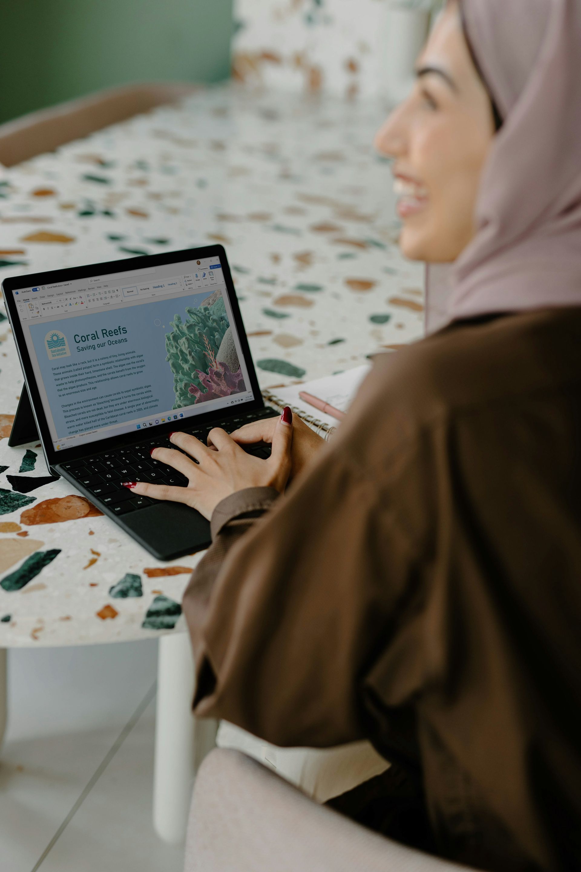 Woman in brown shirt and hijab smiling while working on laptop at a colorful table.