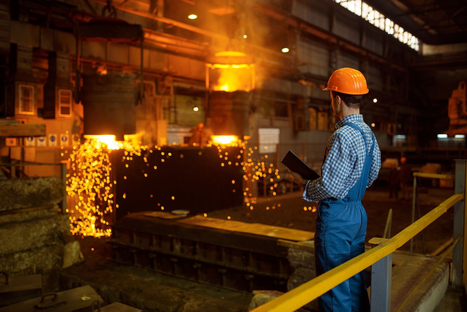 A rear view of a master at a large furnace factory with liquid metal pouring in the background.